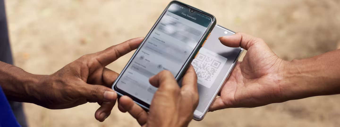 A close-up of two people's hands holding smartphones outdoors. One phone displays a "Transaction Data" form, while the other shows a QR code, representing a digital log or transaction in the field.
