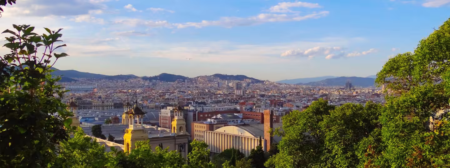 A view of Barcelona from a hilltop with lush greenery framing the skyline and architecture.