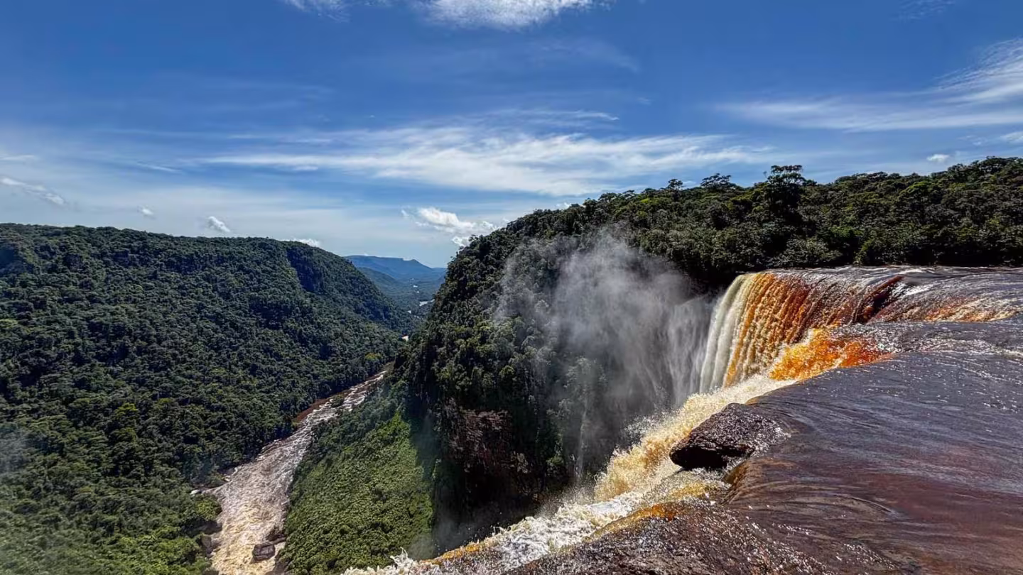 A high-angle view of Kaieteur Falls in Guyana, with amber-colored water cascading into a deep gorge surrounded by a vast rainforest valley.