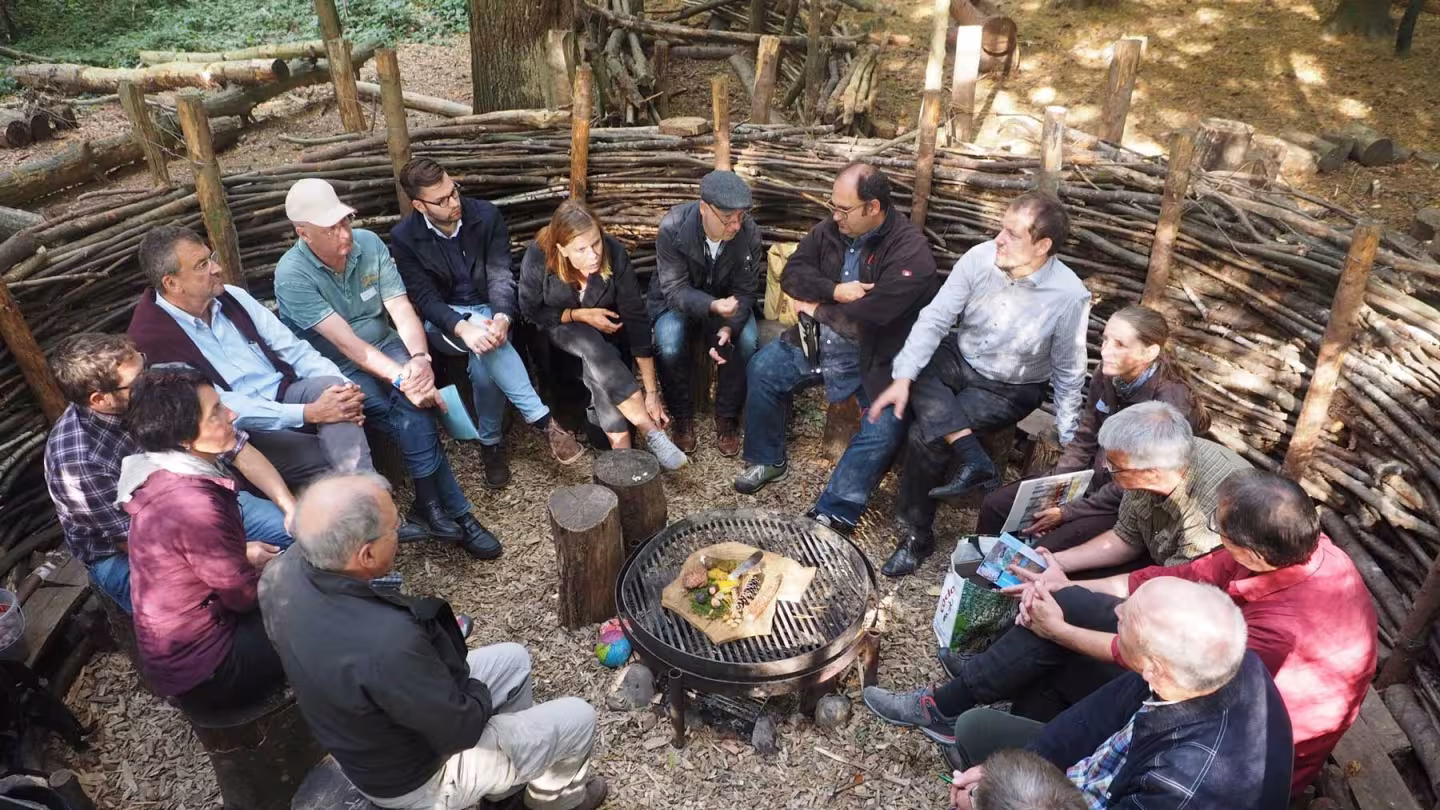 A group of stakeholders sits in a circular outdoor meeting space constructed from woven branches, engaged in discussion around a central fire pit in a forested setting.