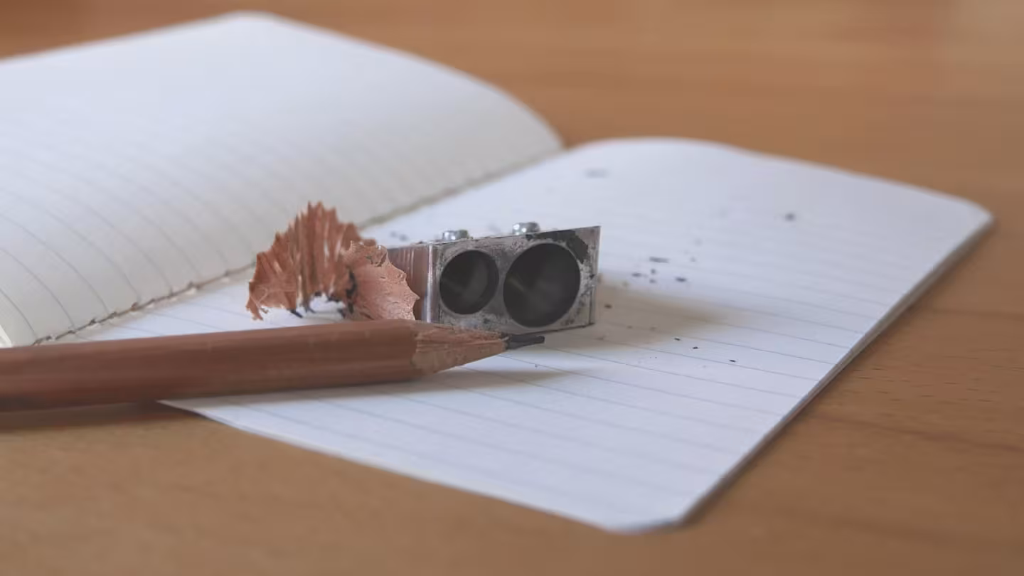 A sharpened wooden pencil, a metal dual-hole sharpener, and a wood shaving resting on an open lined notebook on a wooden desk.