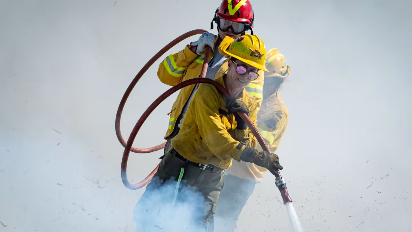 Three firefighters in yellow protective gear work through heavy smoke while handling a pressurized fire hose. The firefighter in the foreground directs a stream of water, while two others stand behind providing support. One firefighter wears a red helmet and protective goggles. Smoke and airborne debris partially obscure the background, emphasizing active wildfire suppression efforts.
