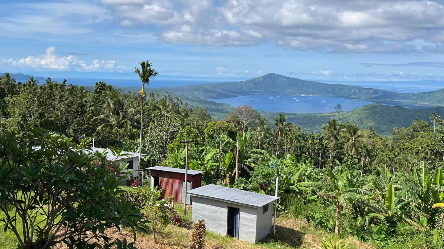 High-angle view of small, metal-roofed buildings surrounded by dense tropical forestry overlooking a bay and volcanic peak in Papua New Guinea.