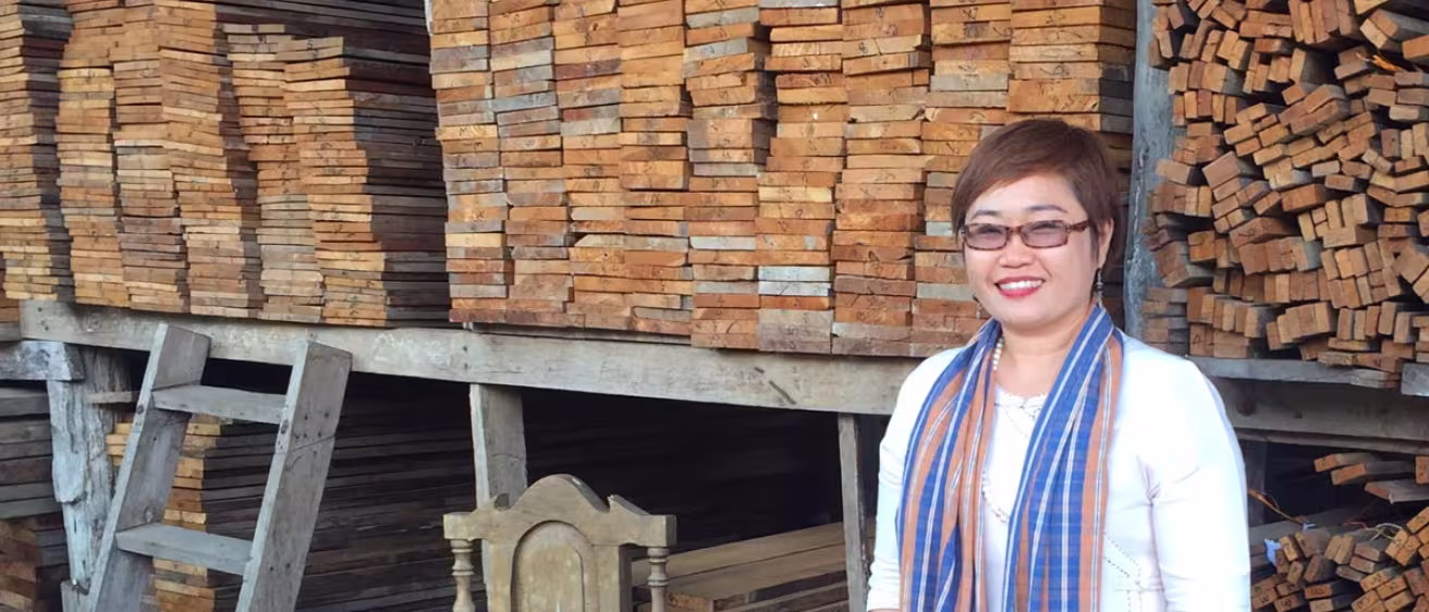 A smiling woman stands in front of large, organized stacks of reclaimed timber planks in a warehouse.