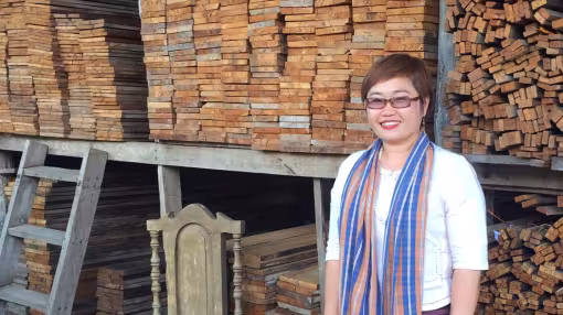 A smiling woman stands in front of large, organized stacks of reclaimed timber planks in a warehouse.