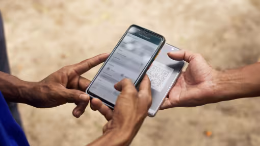 A close-up of two people's hands holding smartphones outdoors. One phone displays a "Transaction Data" form, while the other shows a QR code, representing a digital log or transaction in the field.