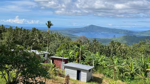 High-angle view of small, metal-roofed buildings surrounded by dense tropical forestry overlooking a bay and volcanic peak in Papua New Guinea.