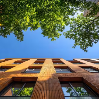 A modern apartment building with a warm wood facade is led amongst lush green trees against a vibrant blue sky.