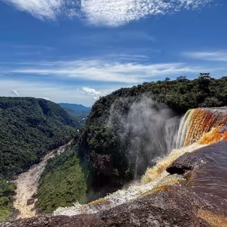 A high-angle view of Kaieteur Falls in Guyana, with amber-colored water cascading into a deep gorge surrounded by a vast rainforest valley.