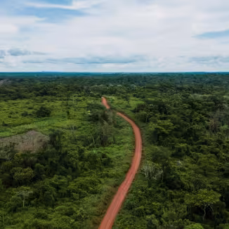 Aerial view of a winding red clay road cutting through a dense green rainforest under a cloudy sky.