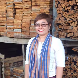 A smiling woman stands in front of large, organized stacks of reclaimed timber planks in a warehouse.