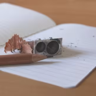 A sharpened wooden pencil, a metal dual-hole sharpener, and a wood shaving resting on an open lined notebook on a wooden desk.