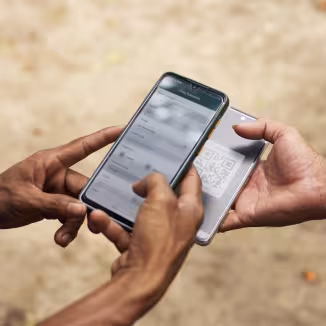 A close-up of two people's hands holding smartphones outdoors. One phone displays a "Transaction Data" form, while the other shows a QR code, representing a digital log or transaction in the field.