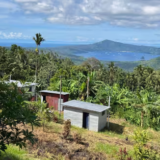 High-angle view of small, metal-roofed buildings surrounded by dense tropical forestry overlooking a bay and volcanic peak in Papua New Guinea.