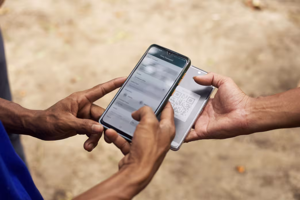 A close-up of two people's hands holding smartphones outdoors. One phone displays a "Transaction Data" form, while the other shows a QR code, representing a digital log or transaction in the field.