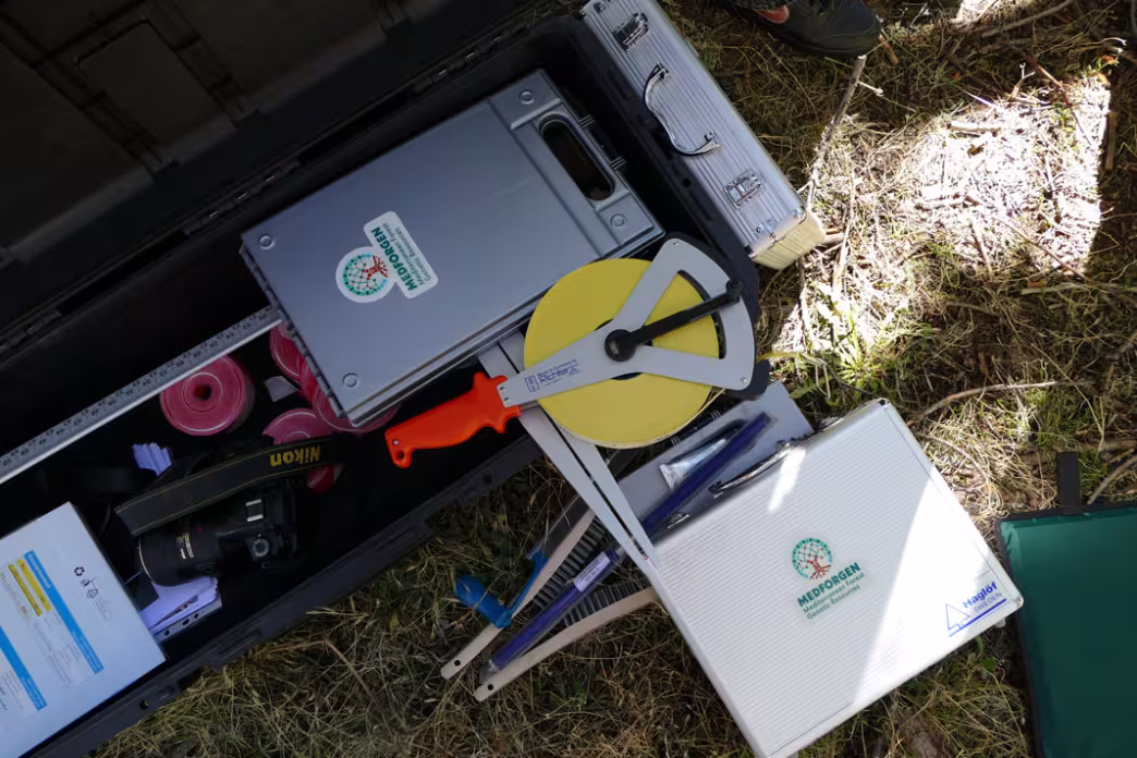 Open equipment case on the grass containing forest inventory tools, including a measuring tape, a calliper, a camera and instrument cases with MEDFORGEN stickers.